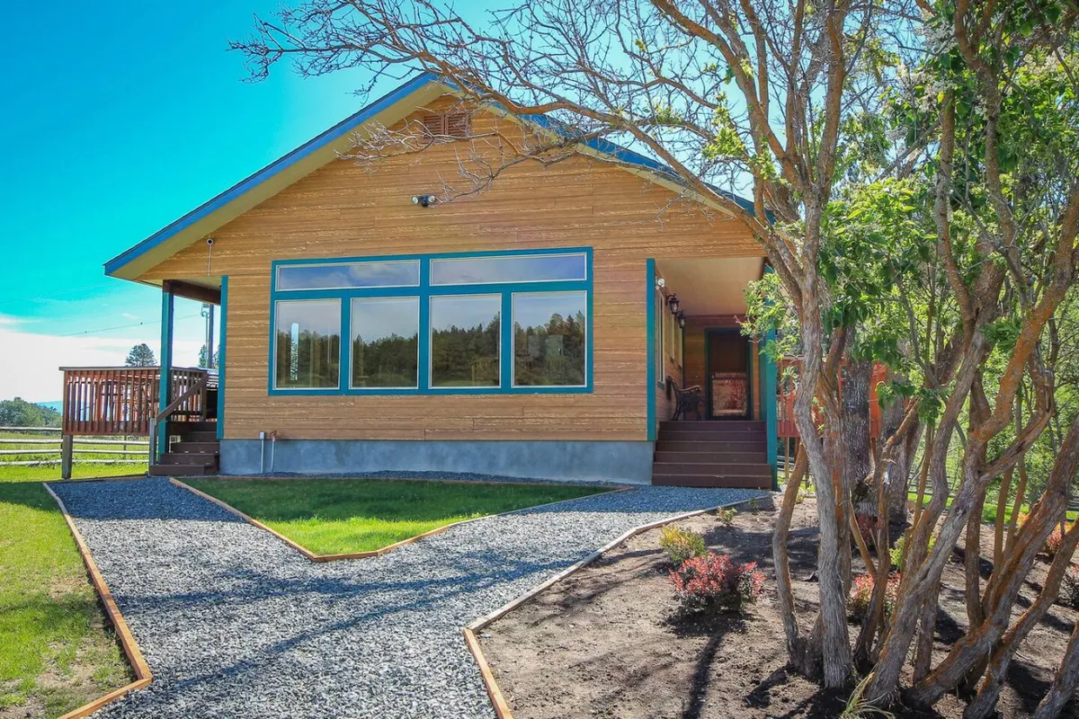 This image features the exterior of the home with large windows, a wooden porch, landscaped pathway, and lush surrounding greenery. Natural light streams into the house, suggesting an inviting setting where indoor spaces connect seamlessly to the peaceful outdoor environment.