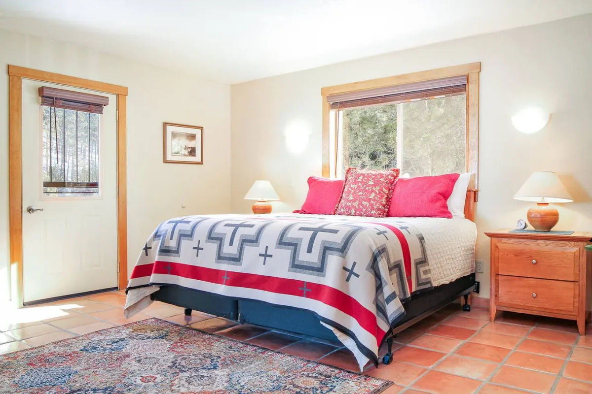 A cozy bedroom with a neatly made bed positioned beside a sunlit window, a nightstand with a lamp, and a door leading to another part of the home. Soft natural light fills the space, creating a restful and inviting atmosphere.