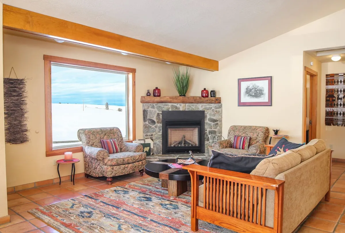 A cozy open-concept living room featuring a gas fireplace as its focal point, surrounded by plush seating, a large area rug, and generous windows that offer sweeping views of the ranch and mountains beyond. The arrangement invites relaxation and conversation, with natural light flooding the space.