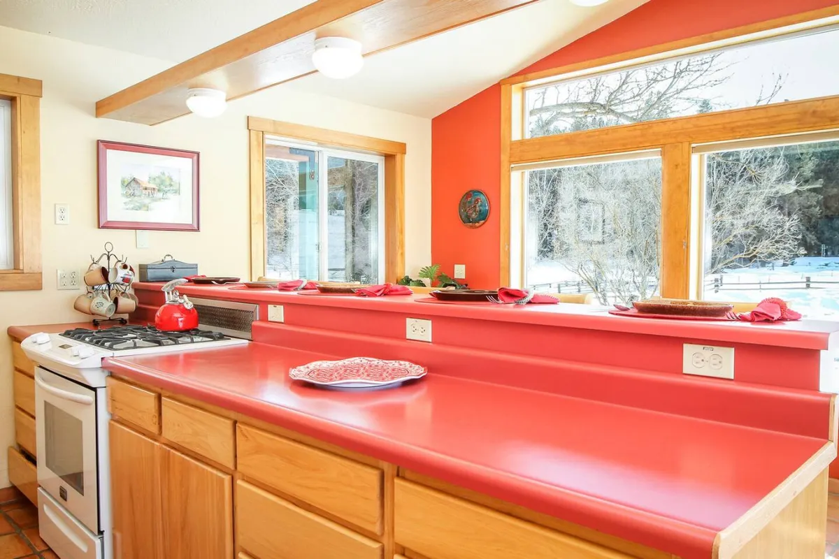 A cheerful kitchen scene with striking red countertops, large windows letting in plenty of light, and wooden cabinetry. Decorative items and ample counter space suggest a lively, functional hub for cooking, entertaining, and interacting with guests or family.