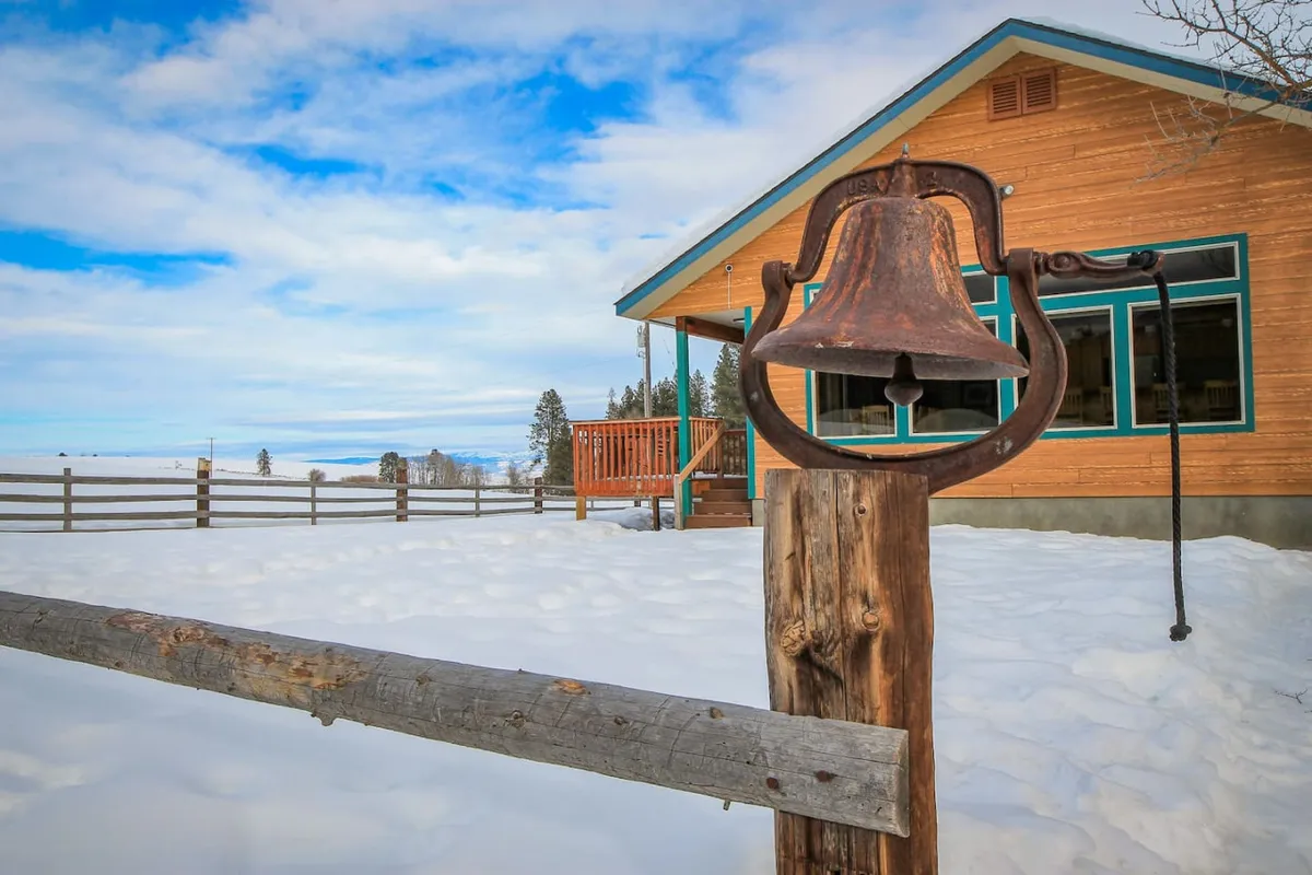 The image showcases the exterior of a welcoming single-family home set against a snow-covered landscape, with a distinctive rusty bell hanging near a classic wooden fence. The cozy house stands invitingly, offering views of the surrounding ranch and mountains, while the scene hints at winter activities like building snowmen and relaxing with warm drinks by the window.
