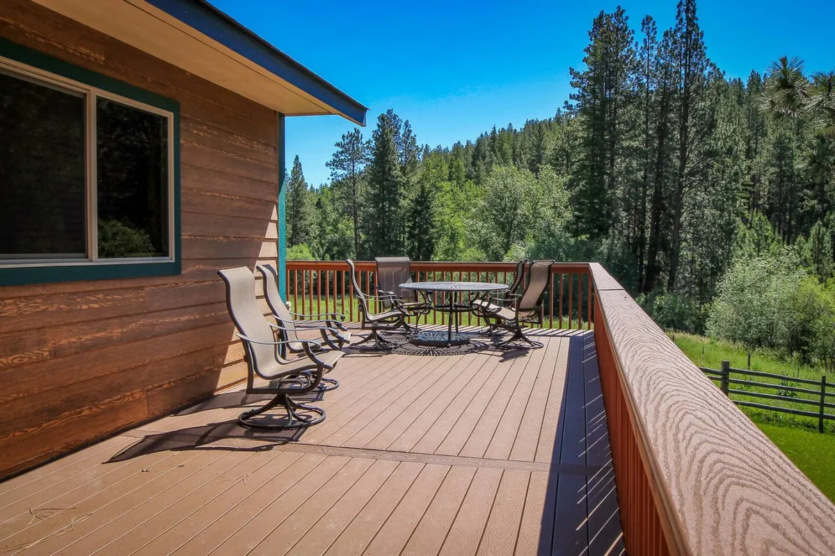 A tranquil shot from the deck, focusing on the railing, outdoor seating, and a table, surrounded by lush trees. The image conveys a sense of calm, inviting guests to relax, socialize, or simply enjoy the natural surroundings at any time of day.