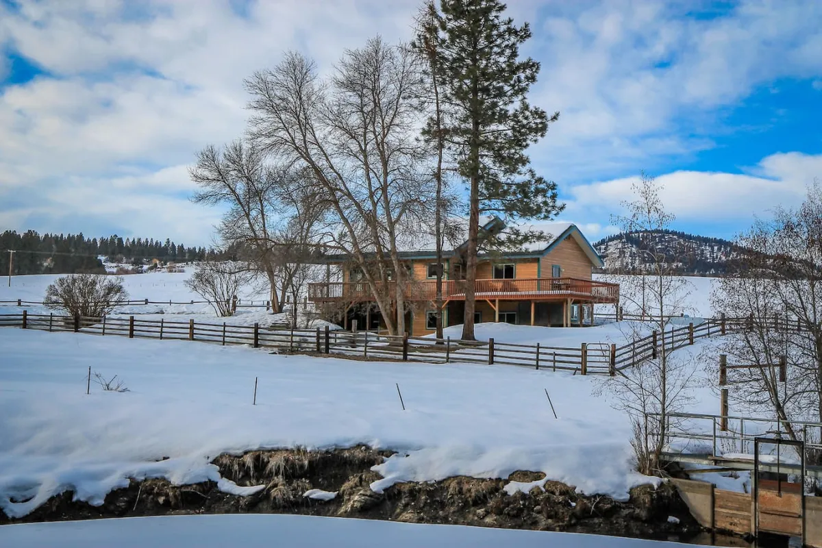 A serene exterior shot of the home set amidst a snow-covered landscape, framed by trees and a rustic fence with distant hills in the background. The peaceful scene evokes the tranquility and privacy of a cozy mountain retreat, ideal for friends or couples looking to relax and enjoy the outdoors together.