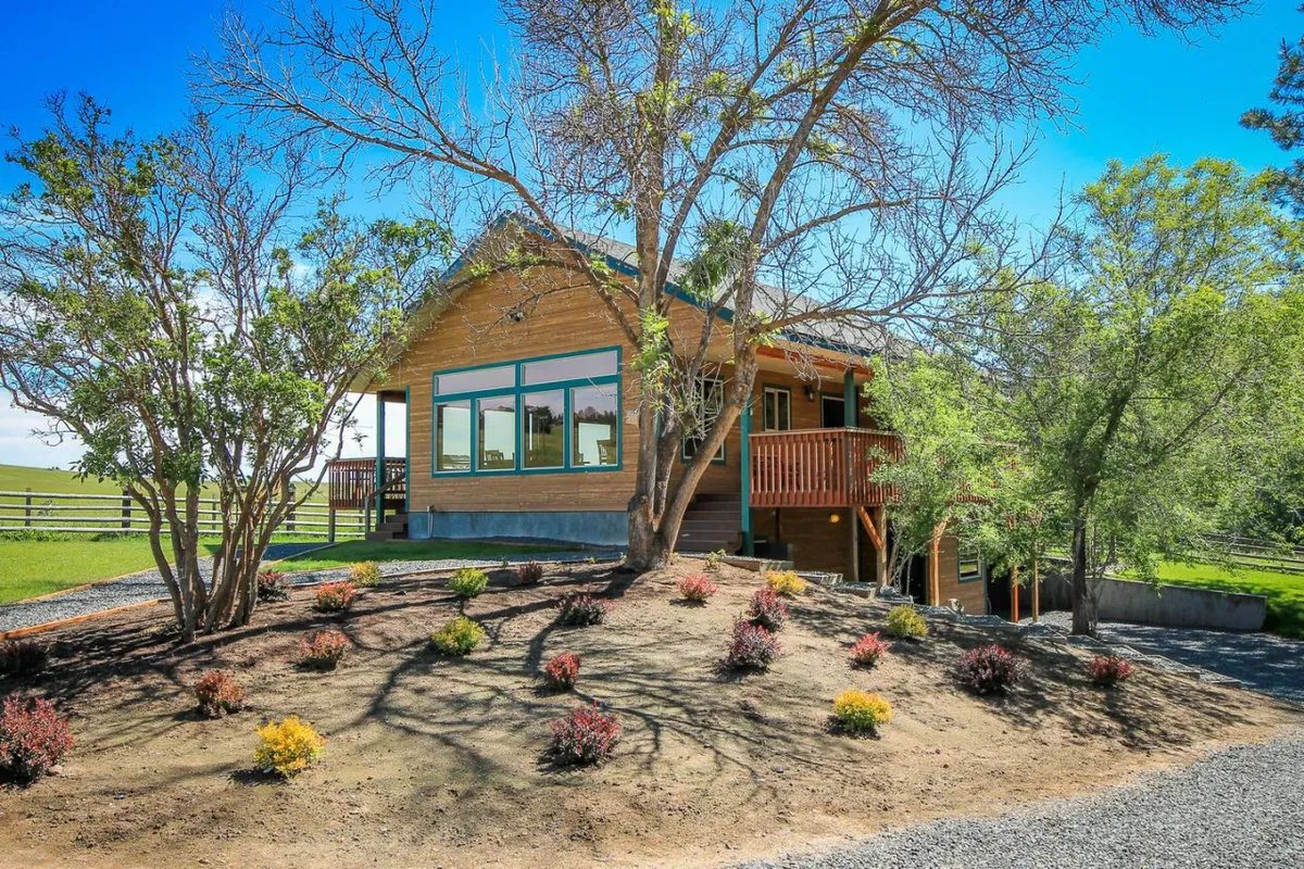 The wooden house sits amid a landscaped garden, framed by tall trees and a front porch with large windows. The scene conveys a peaceful, home-like atmosphere ideal for relaxation or productivity. The porch looks perfect for enjoying morning coffee or an after-work break in nature.