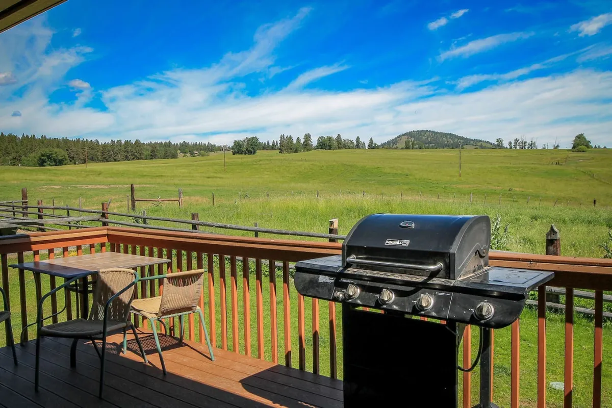 An inviting outdoor dining setup features a grill and a large table set against an expansive green field with mountains in the distance. The scene exudes a relaxed vibe, perfect for al fresco dining, morning coffee, or sunset gatherings, with plenty of space for friends and family to gather.