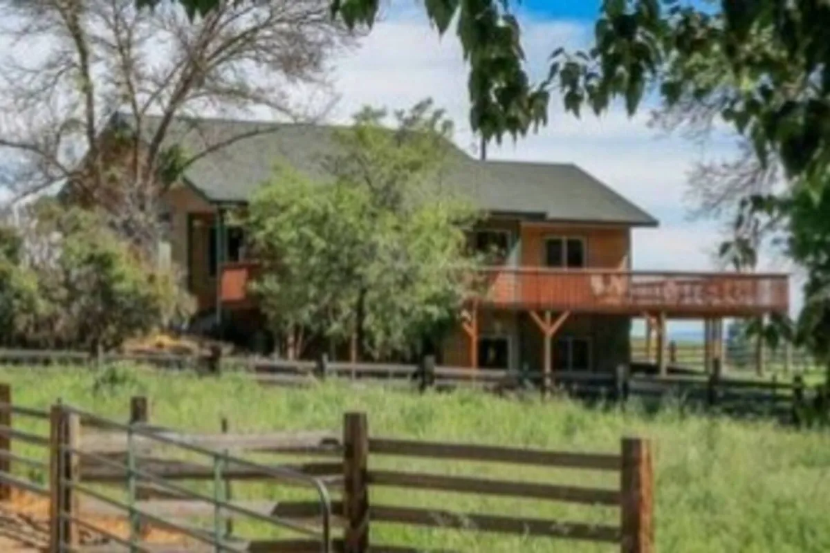 A peaceful exterior shot showcasing the house’s wrap-around deck, surrounded by trees and a wooden fence. The deck is clearly visible, offering a sense of spaciousness and opportunities for outdoor relaxation. The setting sun casts a warm glow, highlighting the inviting atmosphere and natural beauty of the ranch.