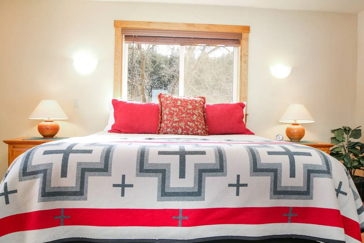 A warm, inviting bedroom with a patterned blanket, vibrant red pillows, bedside lamps, and ample natural light streaming through the window. The space feels cozy and ideal for relaxation.