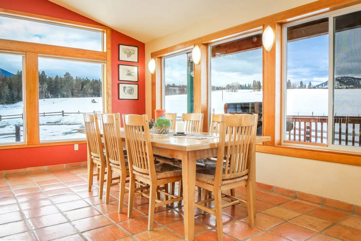 A spacious dining area centered around a large wooden table set for eight, framed by panoramic windows that fill the room with light. Colorful artwork adds vibrancy, creating a space ideal for family meals, celebrations, and social gatherings, all with beautiful views of the snowy landscape outside.