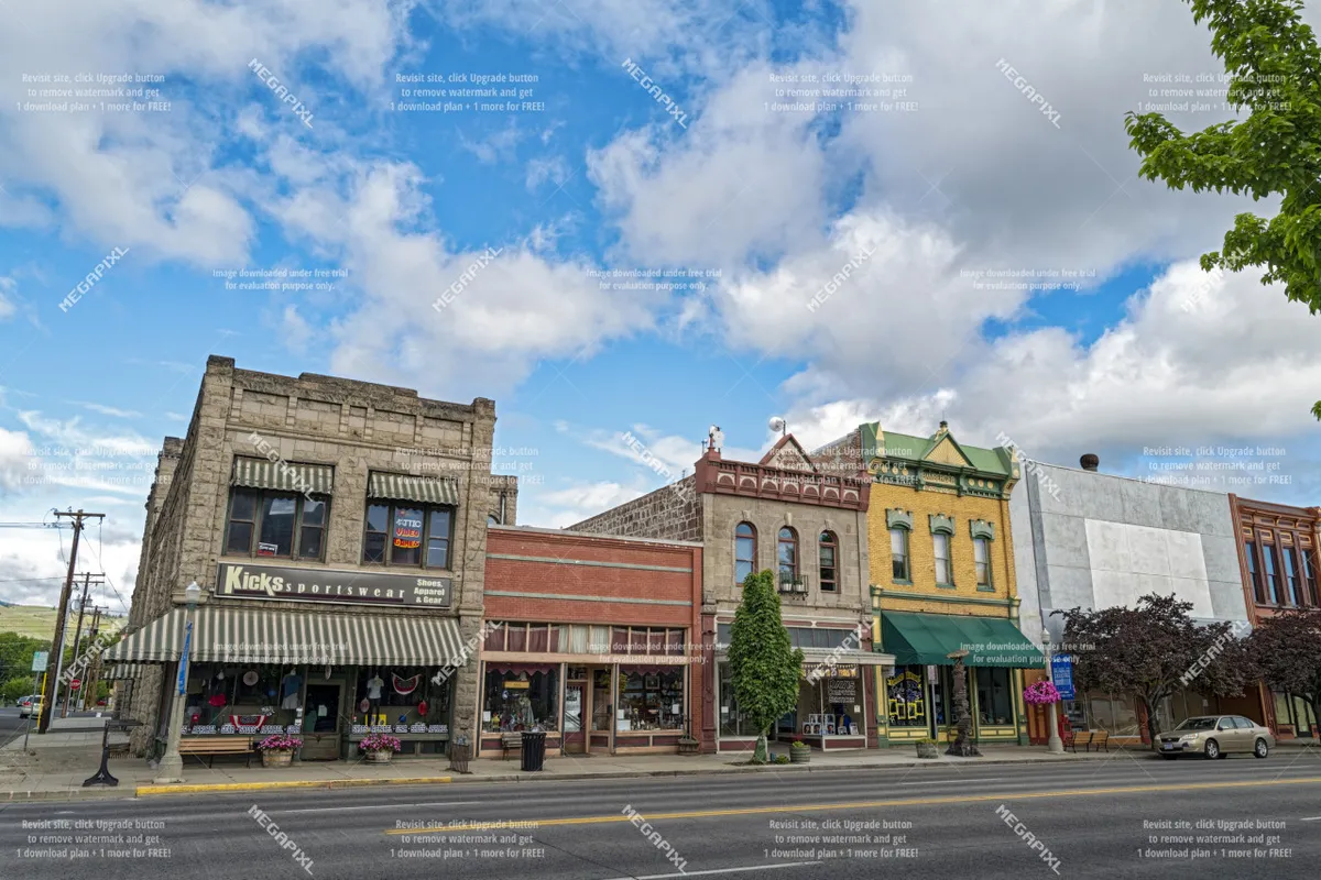 A lively street scene capturing the vibrant storefronts, colorful facades, and leafy trees of downtown Baker City. Visitors are seen strolling, shopping, and enjoying the café culture, reflecting the vibrant local dining and entertainment experiences available nearby.