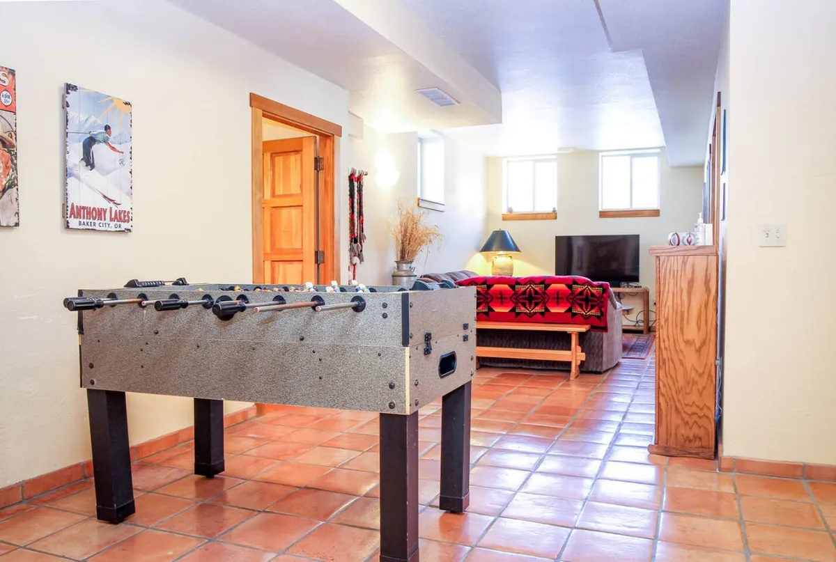 A spacious and vibrant downstairs living room featuring a foosball table at the center, a comfy sofa, a television, decorative wall art, and plenty of natural light through several windows. The arrangement is ideal for playful gatherings, movie nights, and relaxing with friends or family.